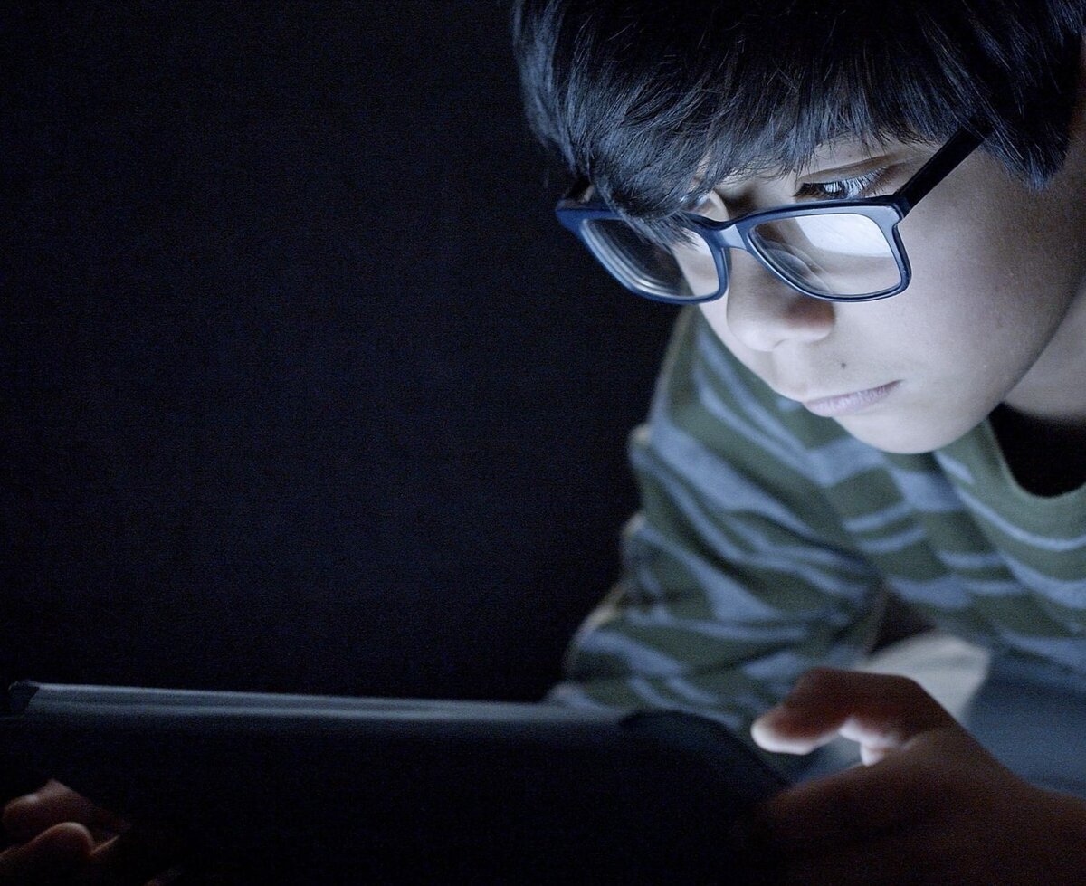 A boy in a darkened room, his face and glasses lit by the glow of a handheld screen