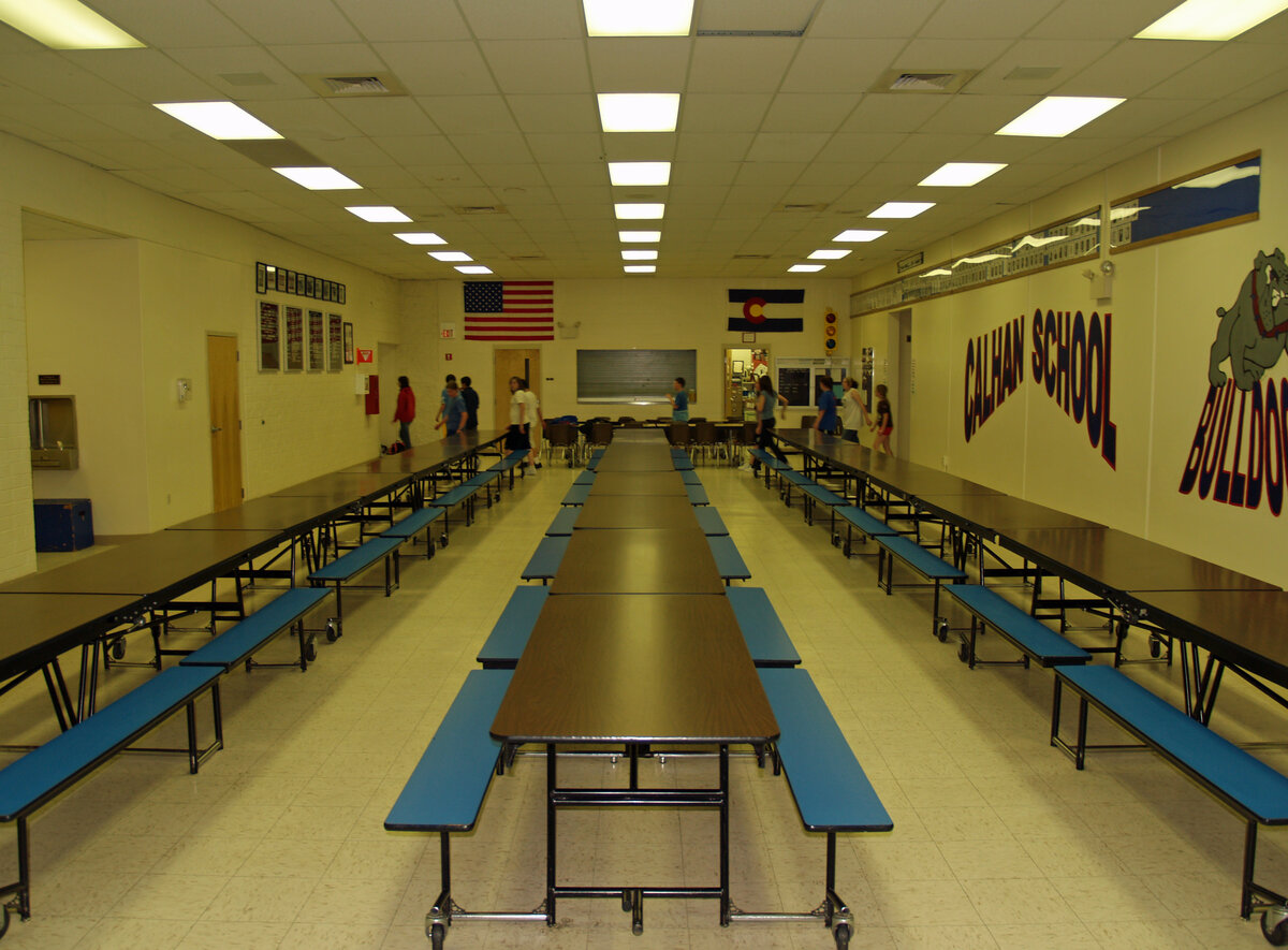 A school cafeteria with long tables under fluorescent lights, the kind of room where eleven forms get signed in nine minutes